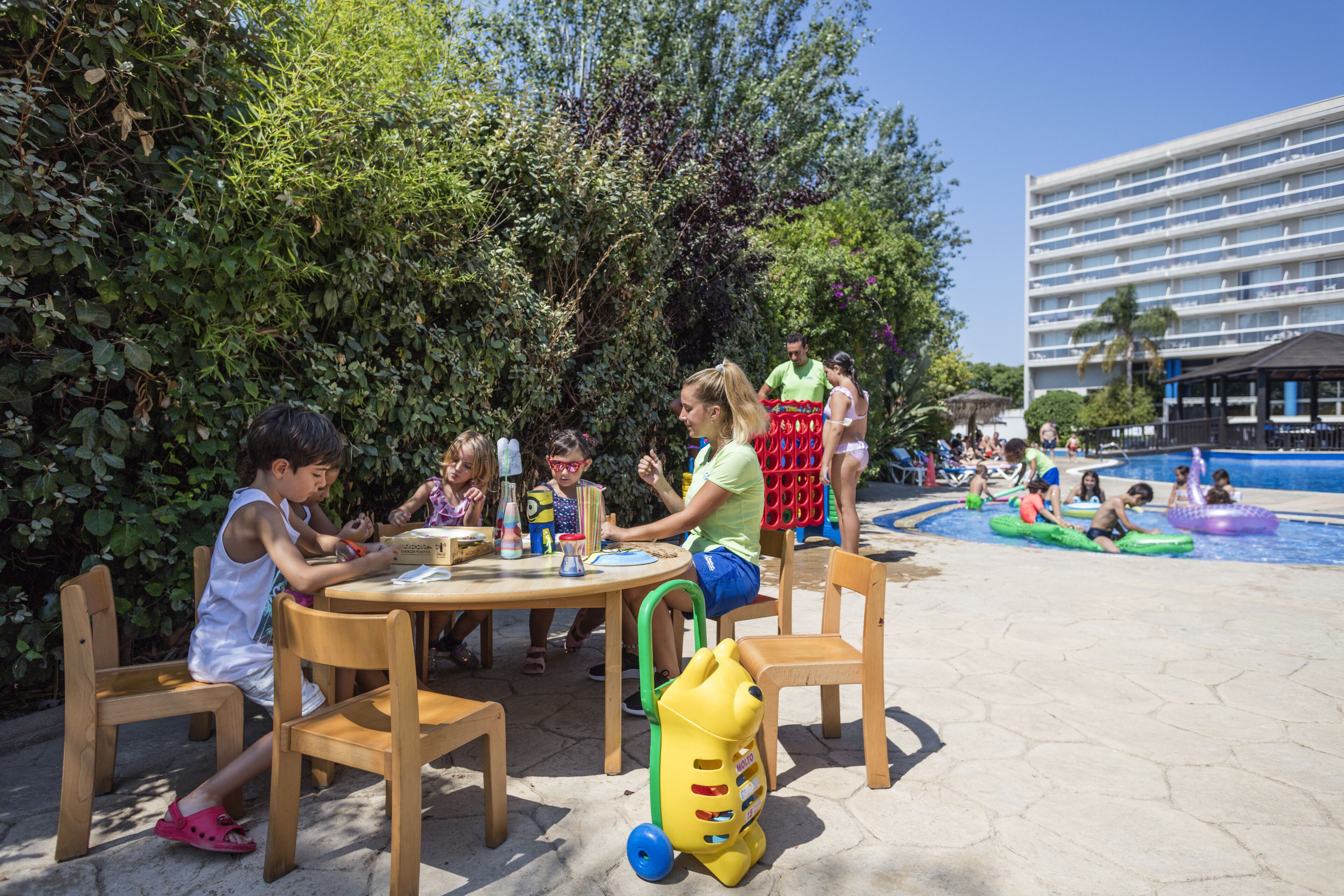 Zona junto a la piscina con niños jugando, una mesa de comedor con sillas y un edificio al fondo.