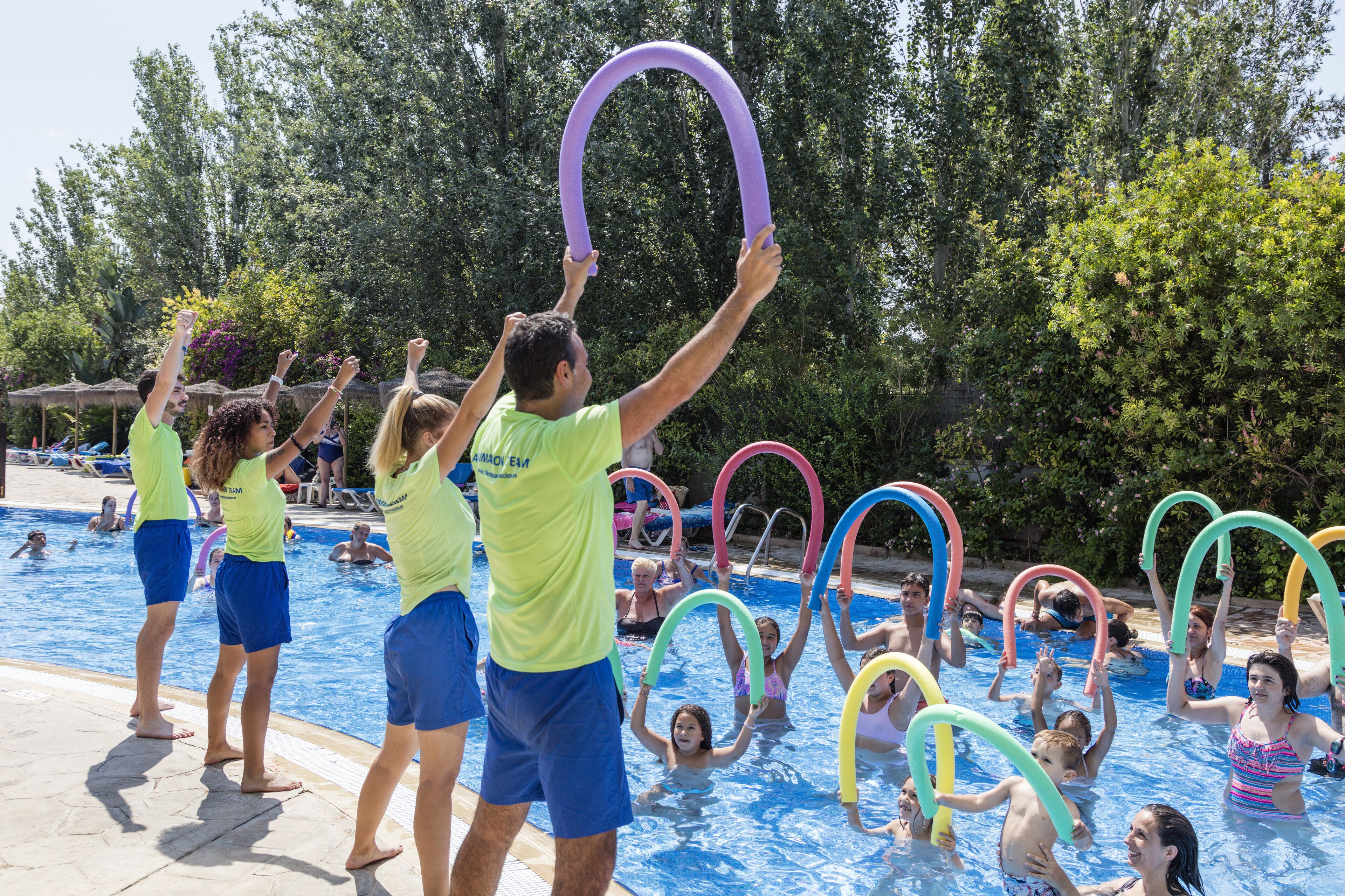 A swimming pool area with people holding colorful rings, surrounded by trees and umbrellas.