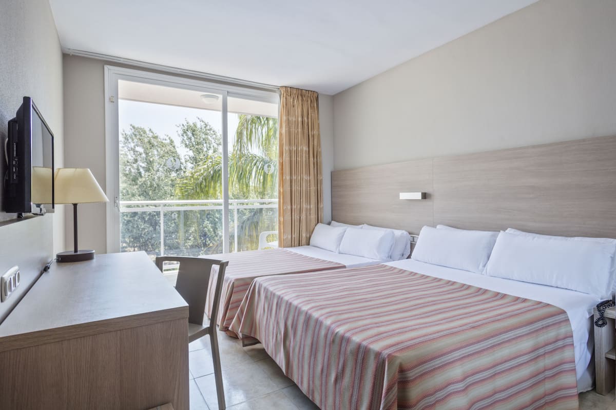 A hotel room with two beds, a desk, a TV, and a balcony with a view of trees.