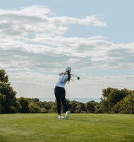Una persona jugando golf en un campo cubierto de césped, con árboles y un cielo despejado al fondo.