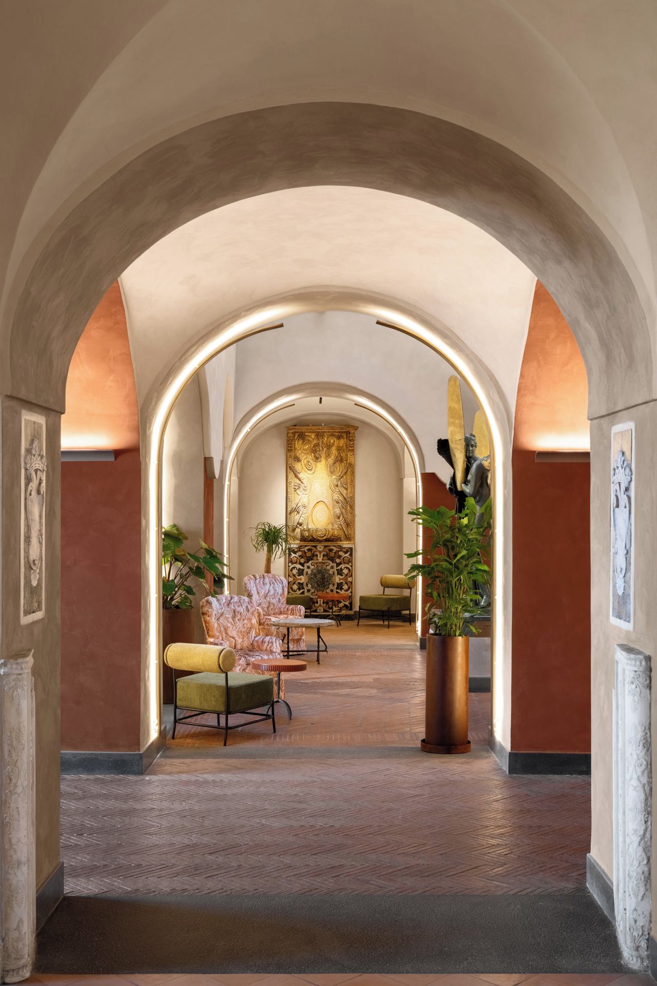 Hall d'albergo con soffitti a volta, una parete decorativa centrale e zone relax con piante.