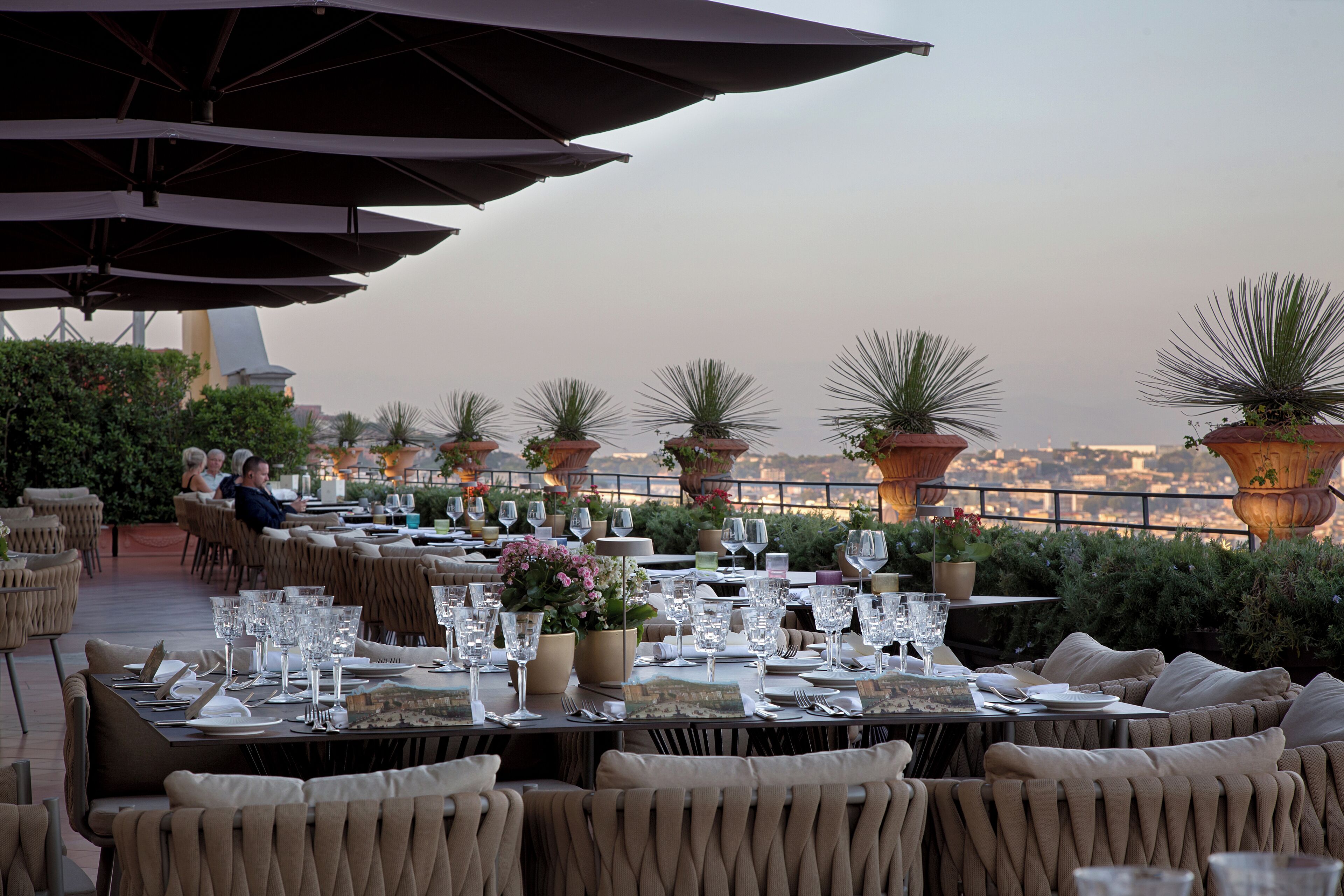An outdoor dining area with tables set for two, surrounded by potted plants and umbrellas.