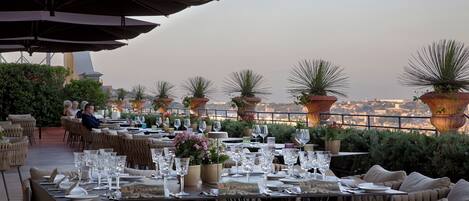 An outdoor dining area with tables set for two, surrounded by potted plants and umbrellas.