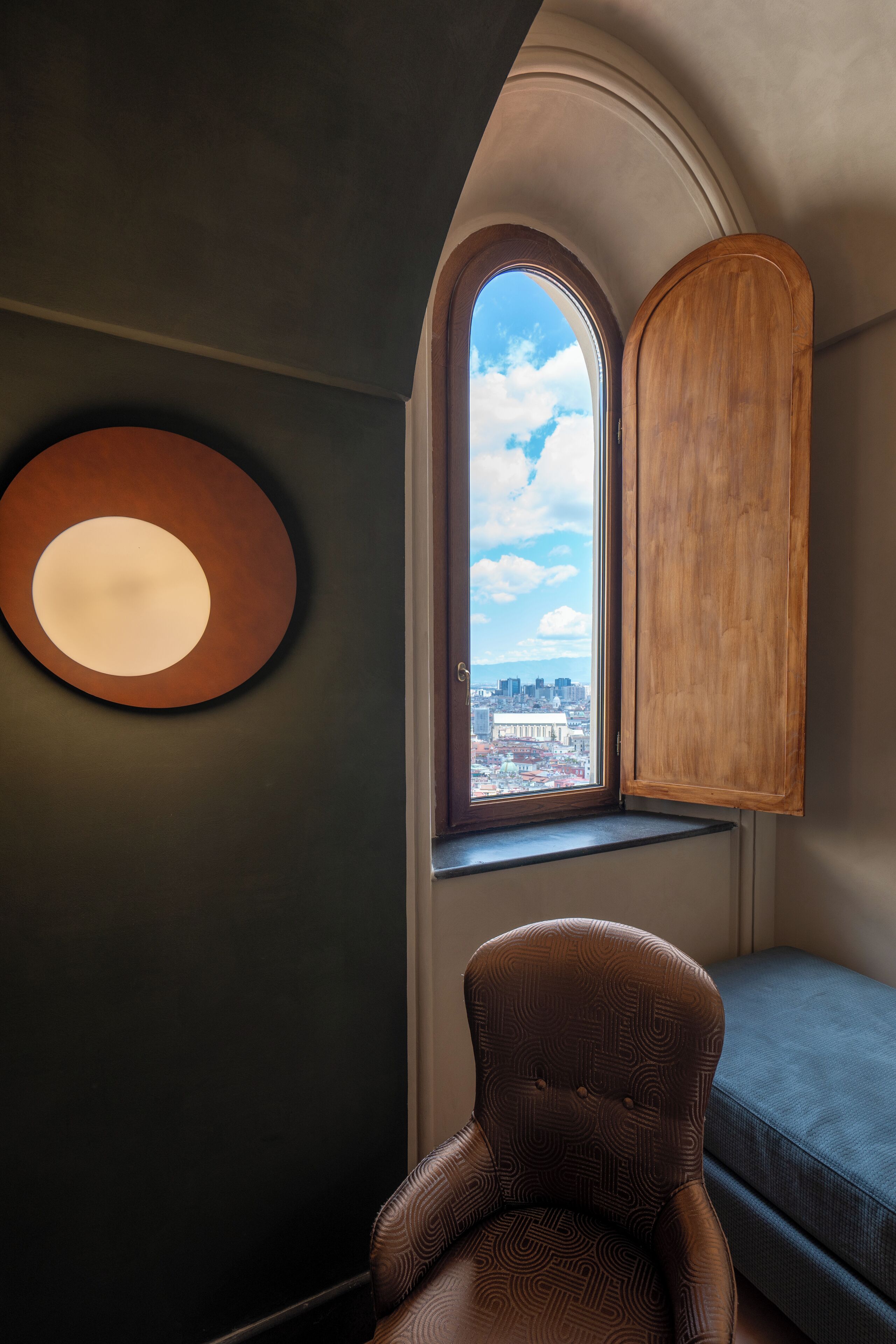 A corner room with a view of a cityscape through a window, a wooden cabinet, a round wall light, and a patterned armchair.
