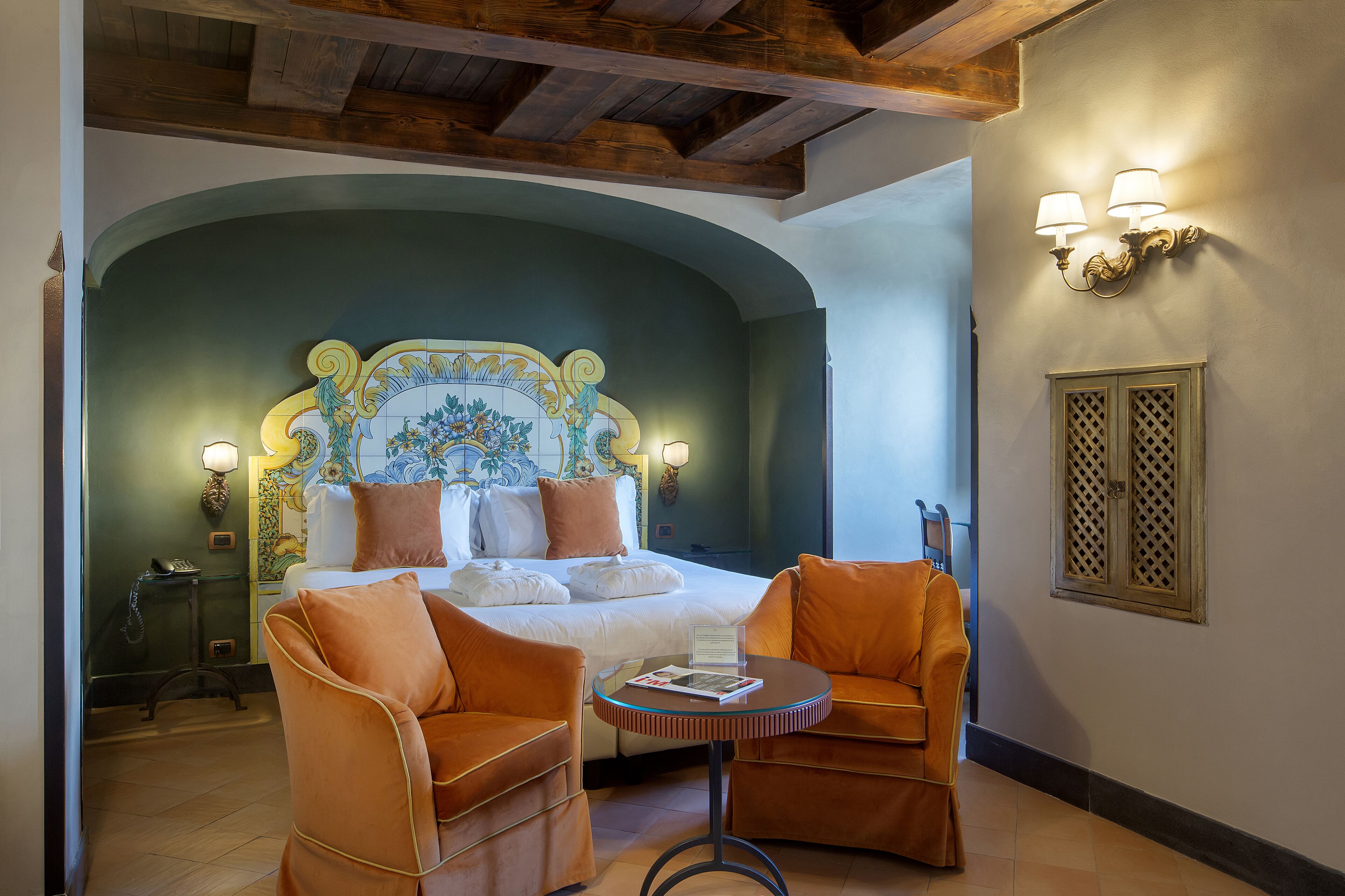 A bedroom with a bed, two orange armchairs, a small round table, and a decorative headboard.