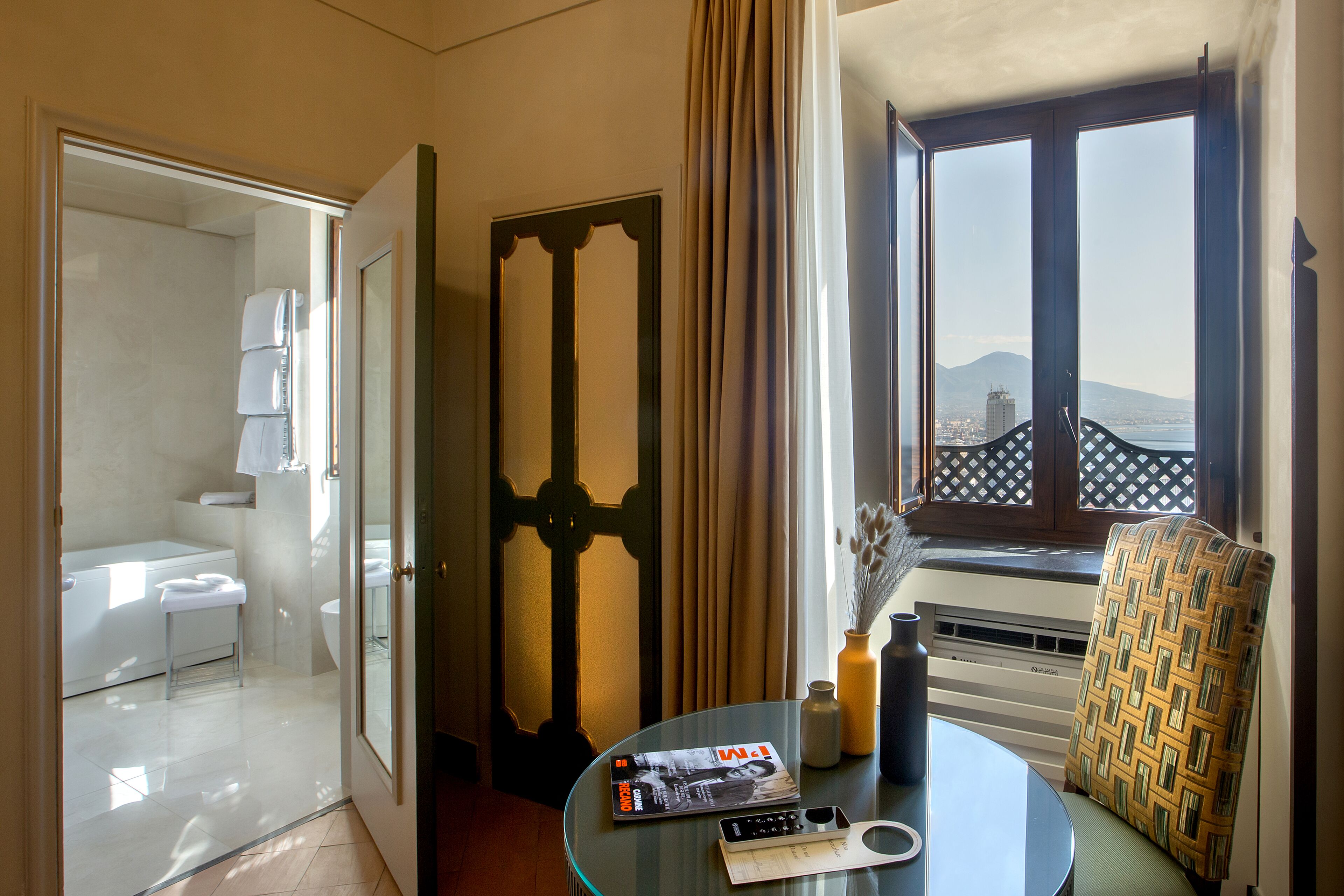 Una camera d'albergo con bagno, zona pranzo con tavolo e sedie, e finestra con vista sulla città.