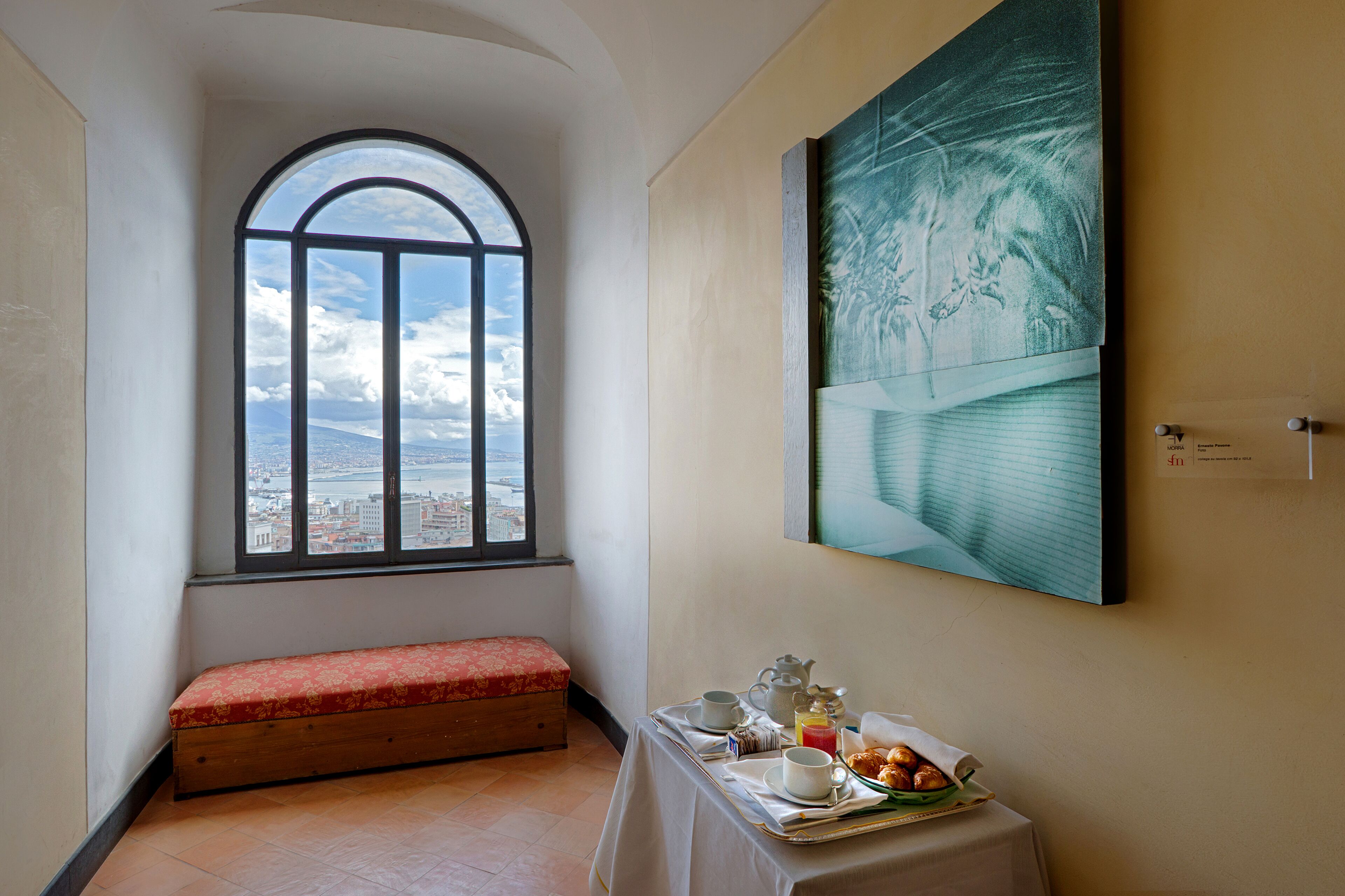 A room with a bench, a table with breakfast items, a large window with a view, and a painting on the wall.
