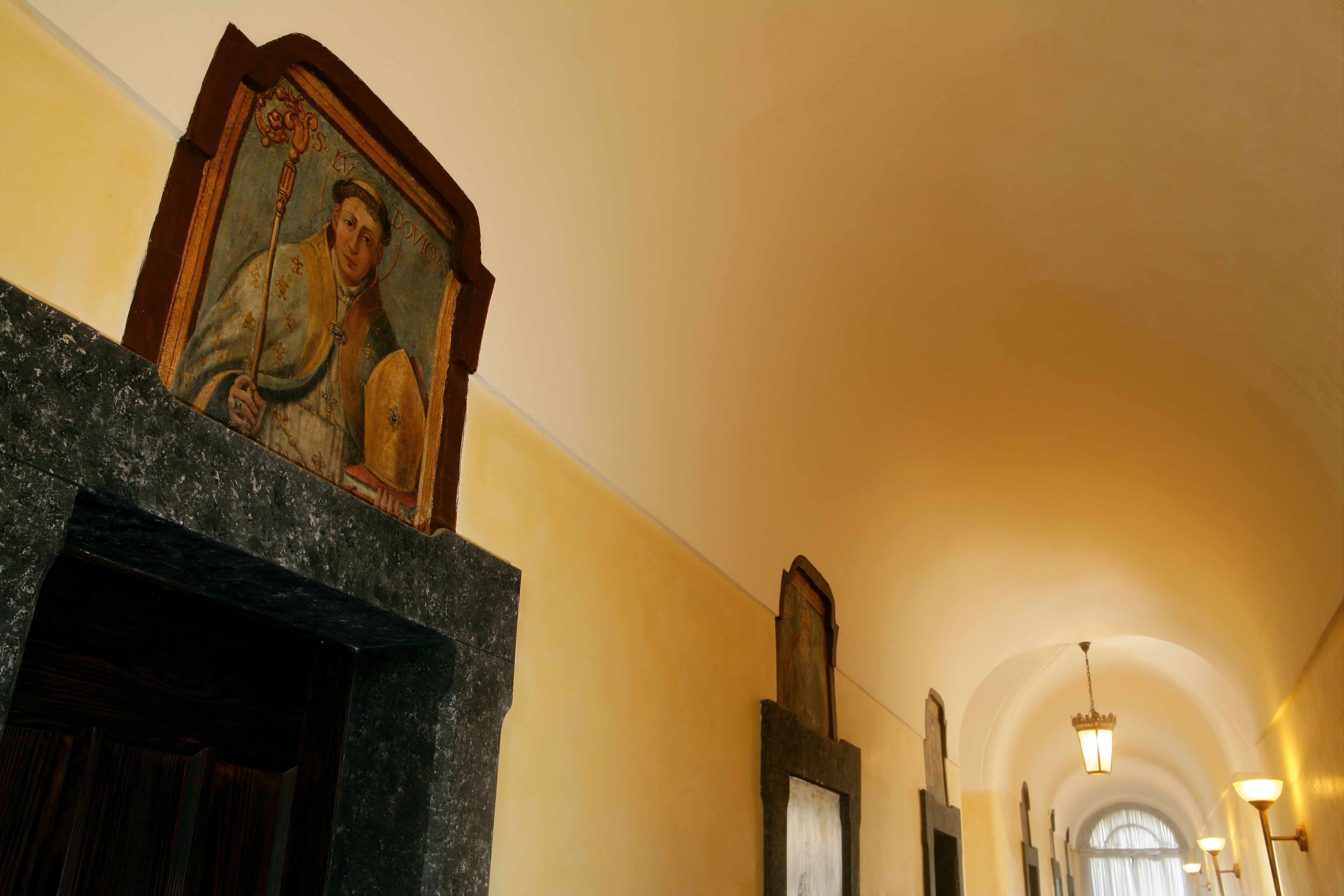 Un couloir avec une cheminée en pierre, un tableau religieux encadré et une porte en bois.
