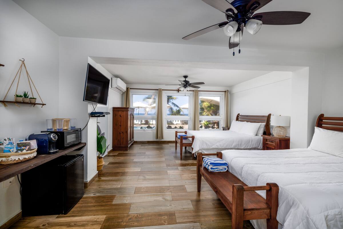A hotel room with two beds, a TV, a ceiling fan, and a view of the ocean through large windows.