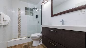 Un baño moderno con cabina de ducha de vidrio, inodoro blanco, tocador de madera oscura y espejo sobre el lavamanos.