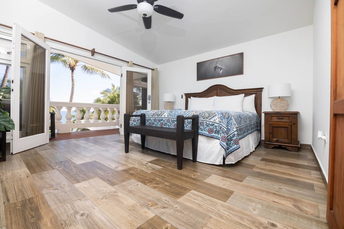 A bedroom with a bed, a bench, a nightstand, a ceiling fan, and a balcony with a view of palm trees.