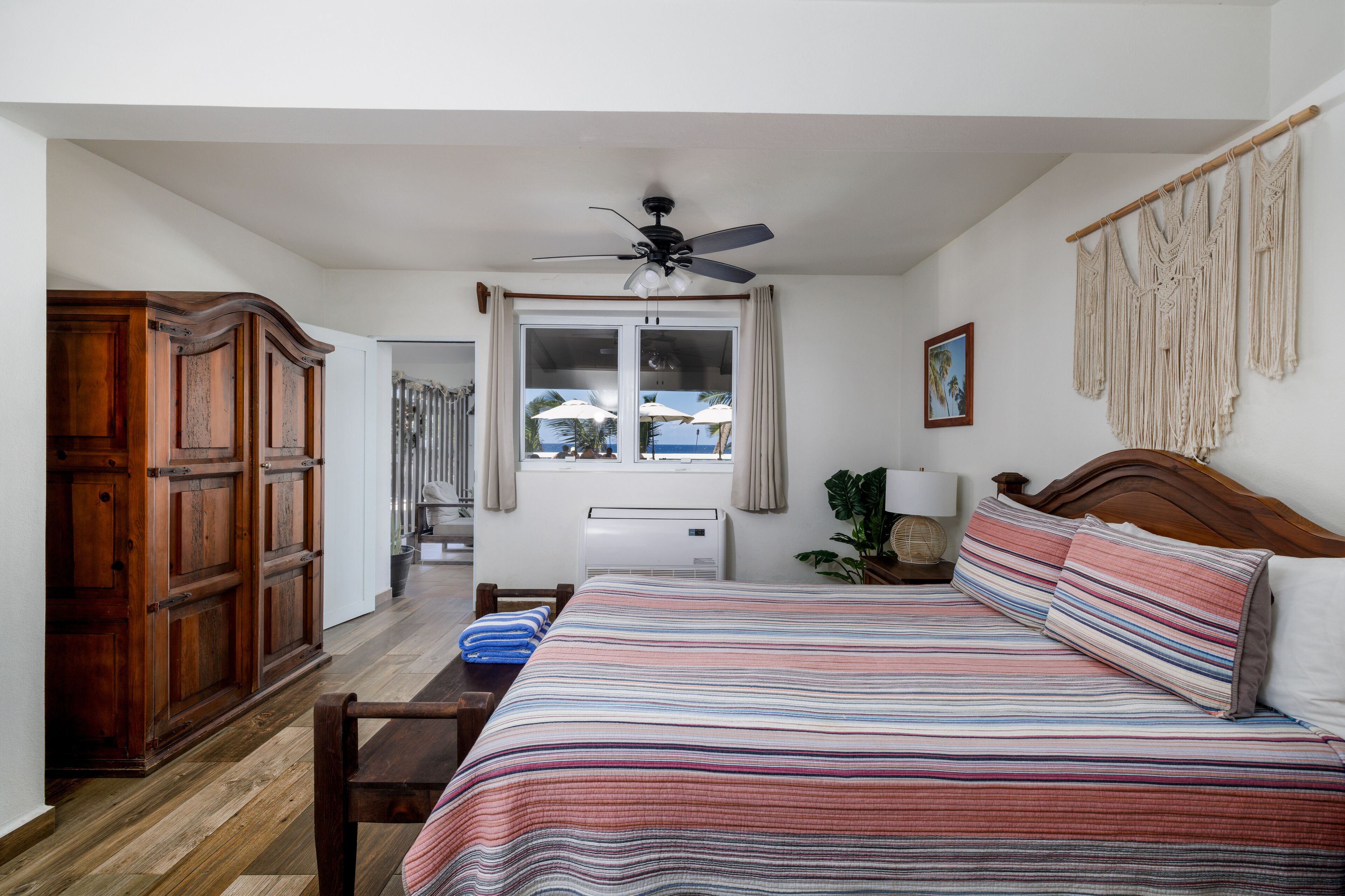 A bedroom with a bed, wooden wardrobe, ceiling fan, window with curtains, and a view of the beach.