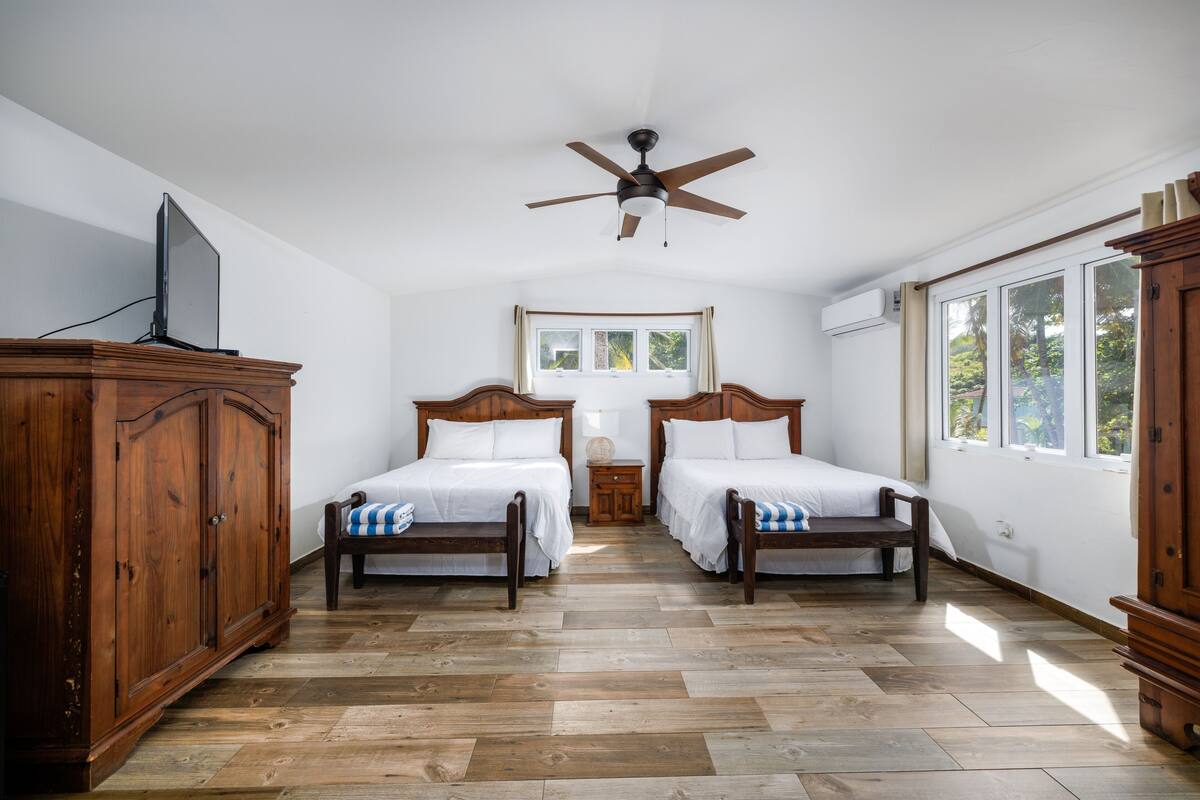 A bedroom with two wooden beds, a ceiling fan, and a large window with a view of greenery.