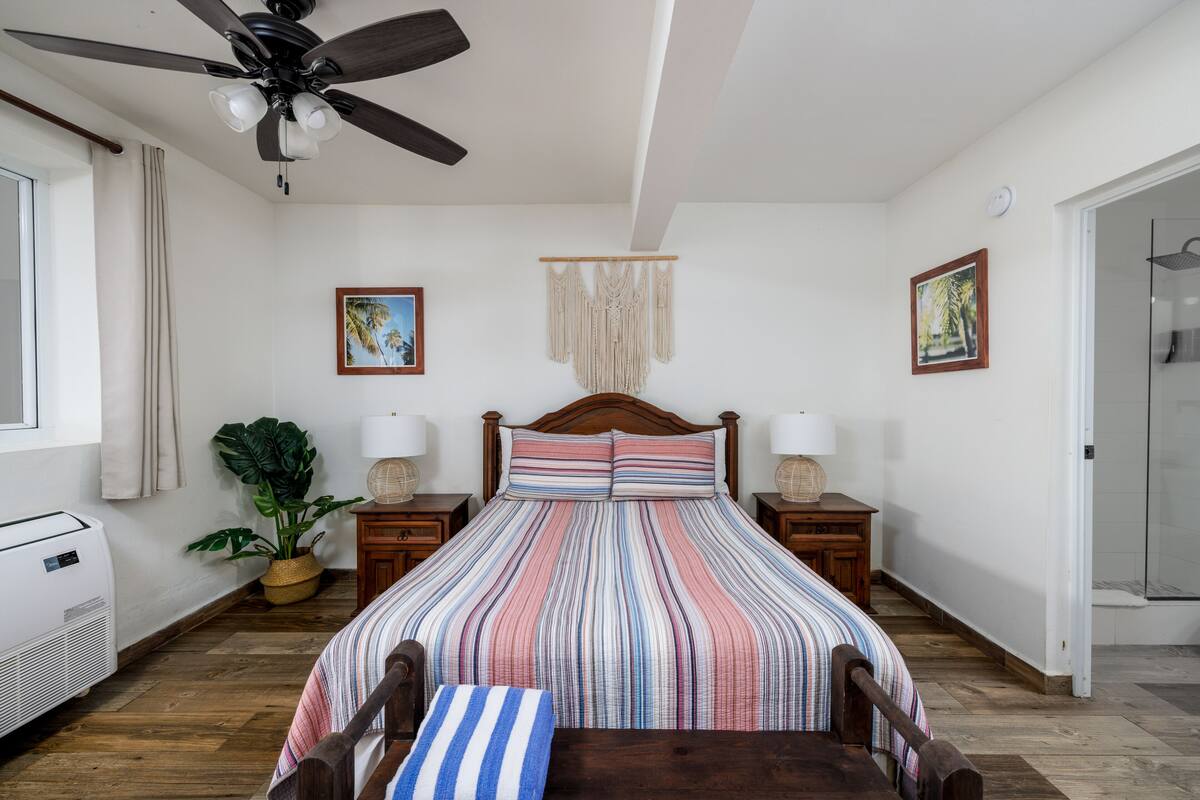 A bedroom with a bed, wooden headboard, bedside tables with lamps, a ceiling fan, a wall-mounted macrame, and a potted plant.