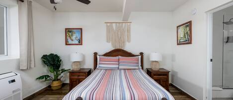 Una camera da letto con un letto, una testiera in legno, comodini con lampade, una ventola a soffitto, un pannello macramè a parete e una pianta in vaso.