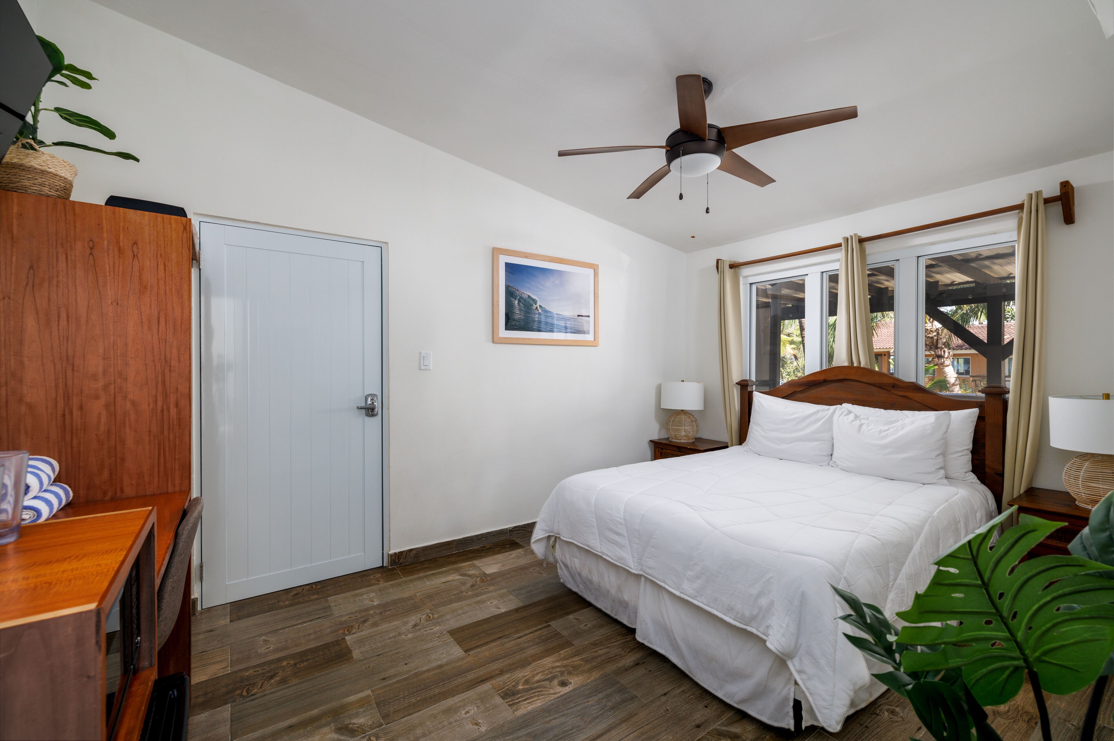 A bedroom with a bed, wooden headboard, bedside tables, a ceiling fan, a window with curtains, a door, and a plant.