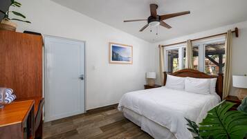 A bedroom with a bed, wooden headboard, bedside tables, a ceiling fan, a window with curtains, a door, and a plant.