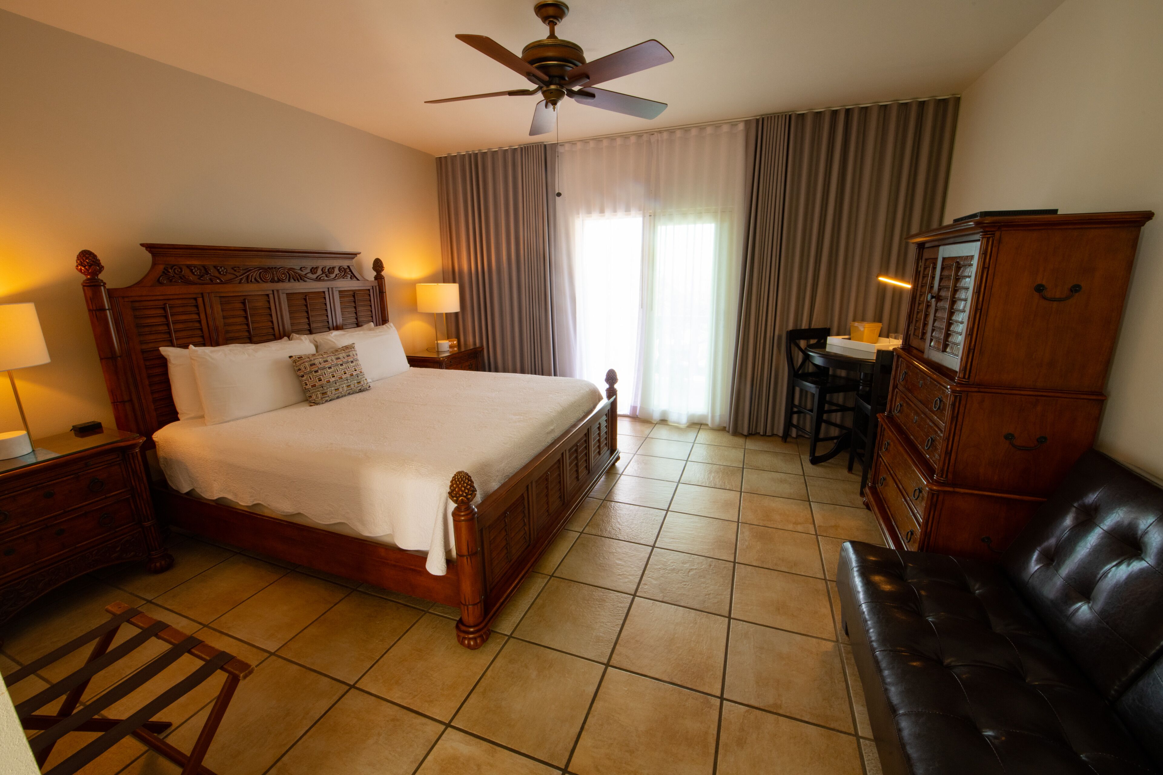 A hotel room with a large bed, a ceiling fan, a desk, a chair, a dresser, and a sofa.