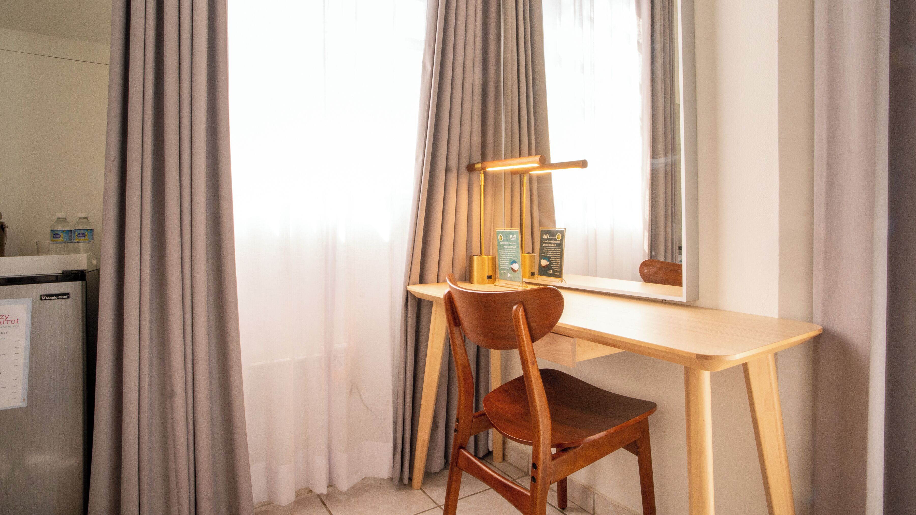 A hotel room with a wooden desk, a chair, a mirror, and a window with curtains.