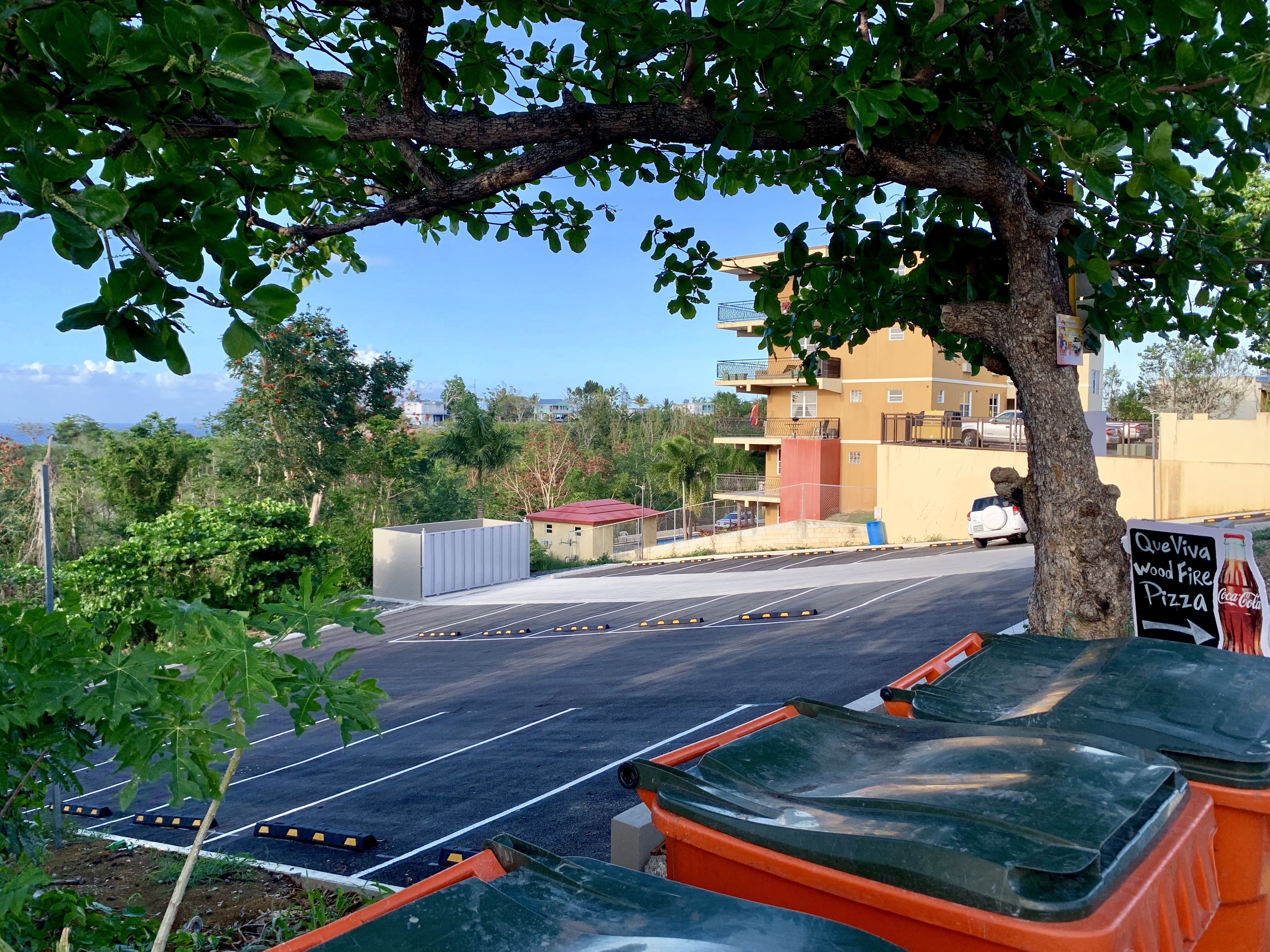 A parking lot with orange trash bins, a tree, and a building with balconies in the background.