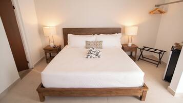 Una cama bien hecha con estructura de madera, dos mesitas de noche con lámparas, un taburete de madera y un tendedero de pared.