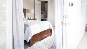 A bedroom with a wooden bed, white curtains, a nightstand, and a lamp. - Lazy Parrot Inn (Rincon)