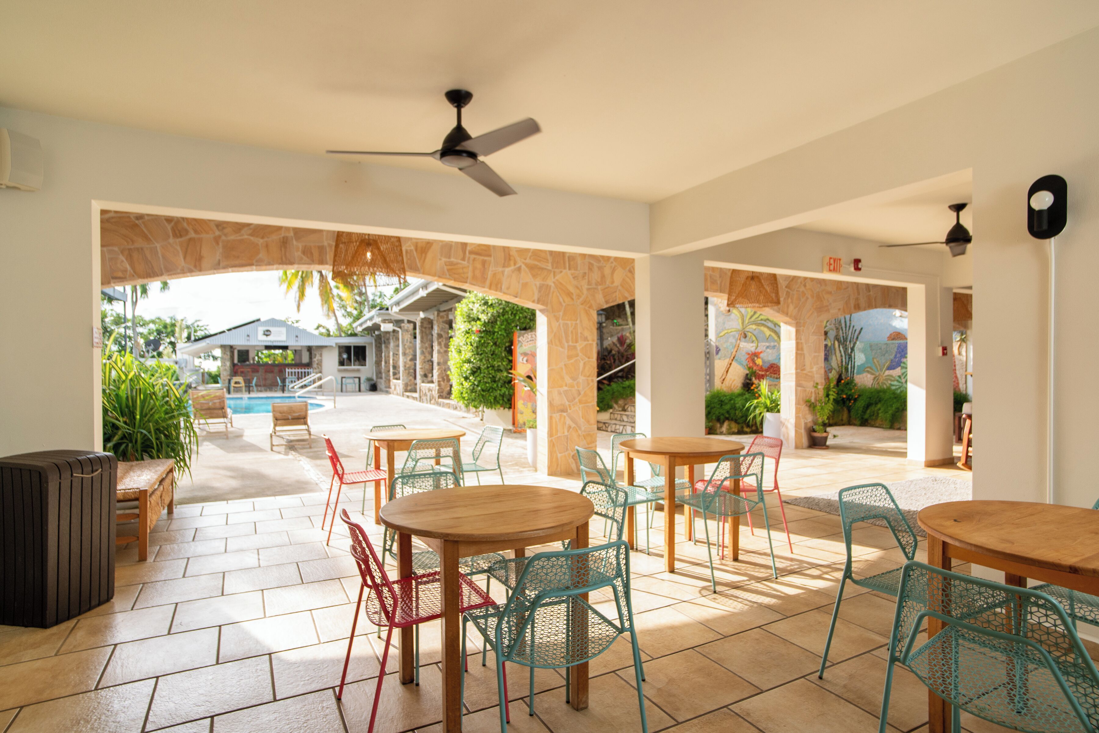A covered outdoor seating area with tables and chairs, a ceiling fan, and a view of a pool and greenery.