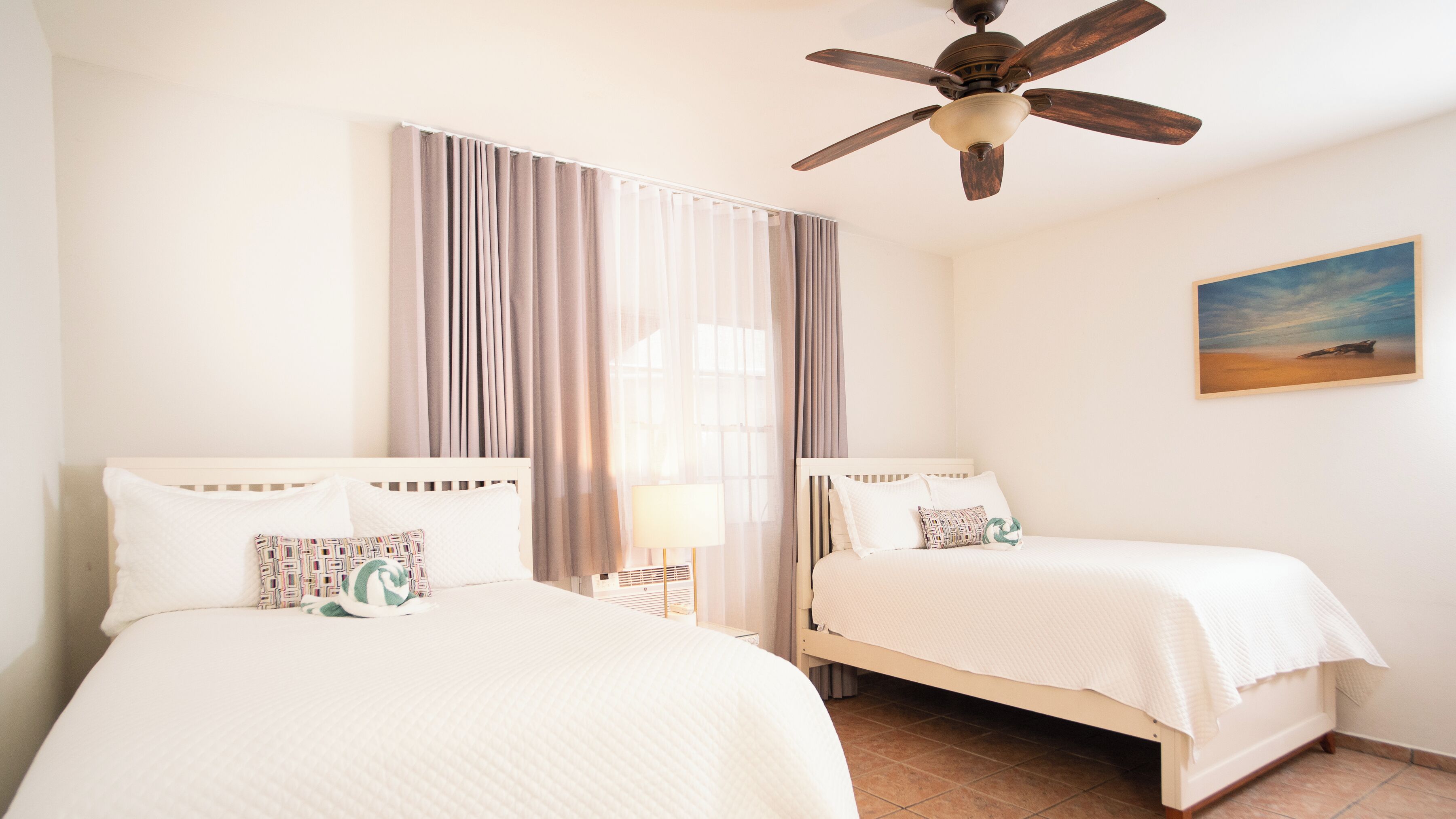 A hotel room with two beds, a ceiling fan, a large window with curtains, and a painting on the wall.