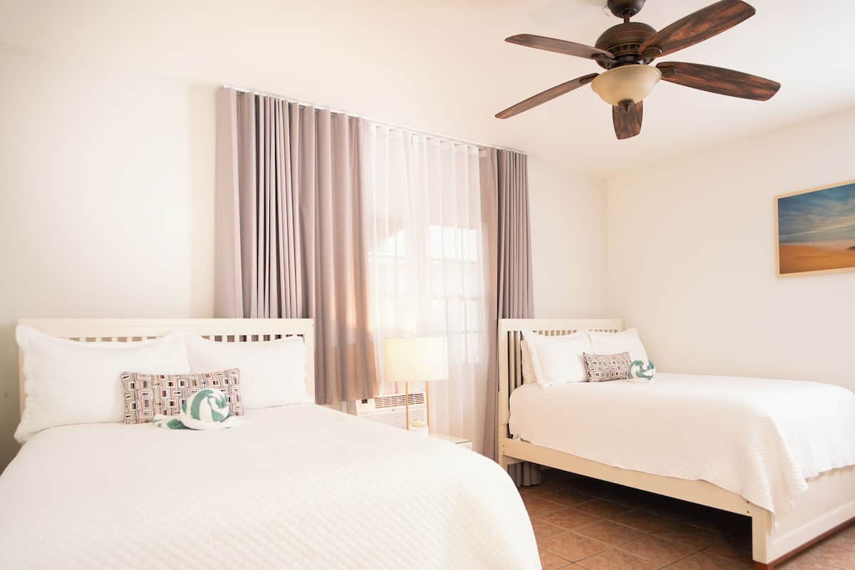 A hotel room with two beds, a ceiling fan, a large window with curtains, and a painting on the wall.