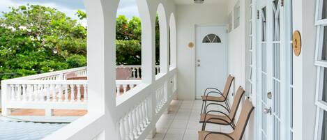 A covered balcony with white railings, a tiled floor, and a row of chairs.