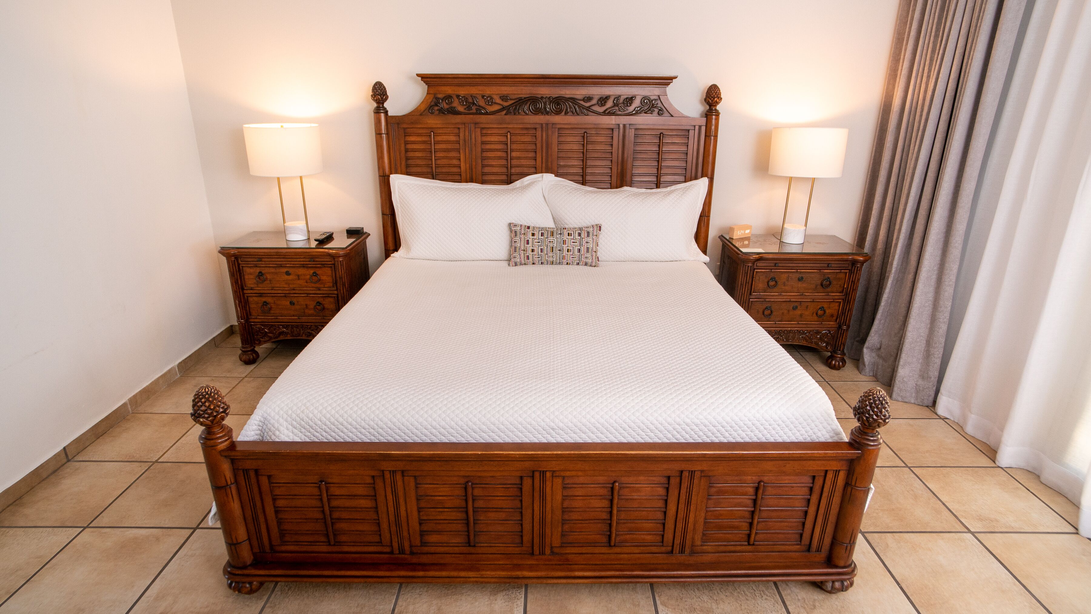 Una cama de madera grande con una colcha blanca acolchada, flanqueada por dos mesitas de noche con lámparas, en una habitación con suelo de baldosas.
