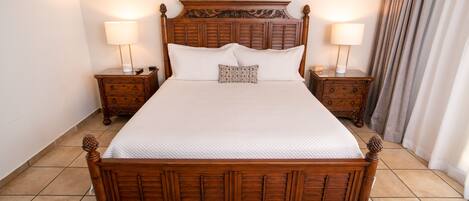A large wooden bed with a quilted white bedspread, flanked by two nightstands with lamps, in a room with tiled flooring.