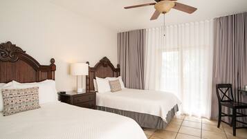 A hotel room with two beds, a nightstand with a lamp, a ceiling fan, and a large window with curtains.