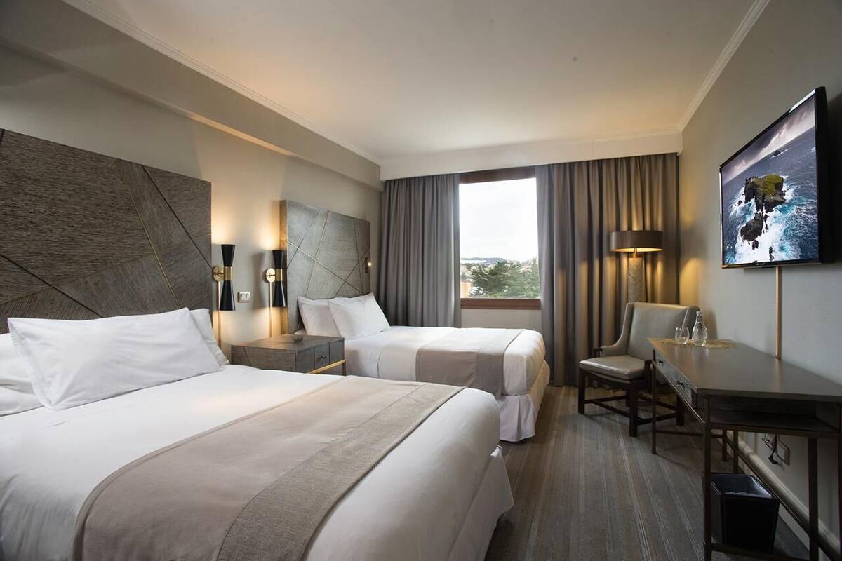 A hotel room with two beds, a desk, a chair, a TV, and a large window with curtains.