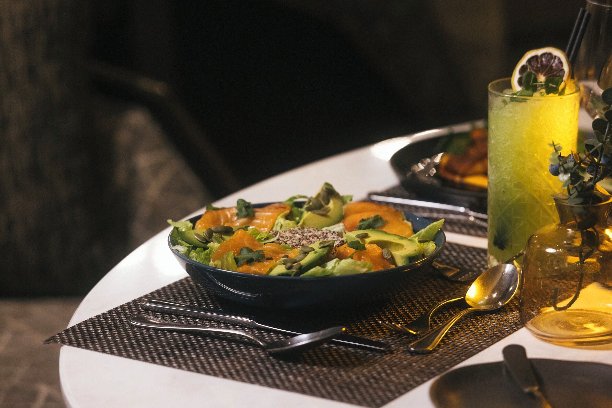 A dining table with a bowl of salad, a glass of drink, and cutlery.