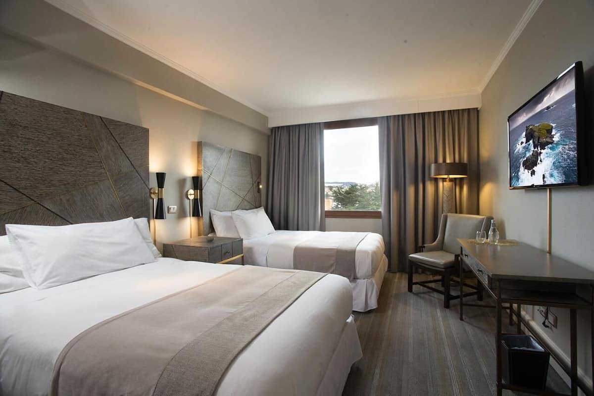 A hotel room with two beds, a desk, a chair, a TV, and a large window with curtains.