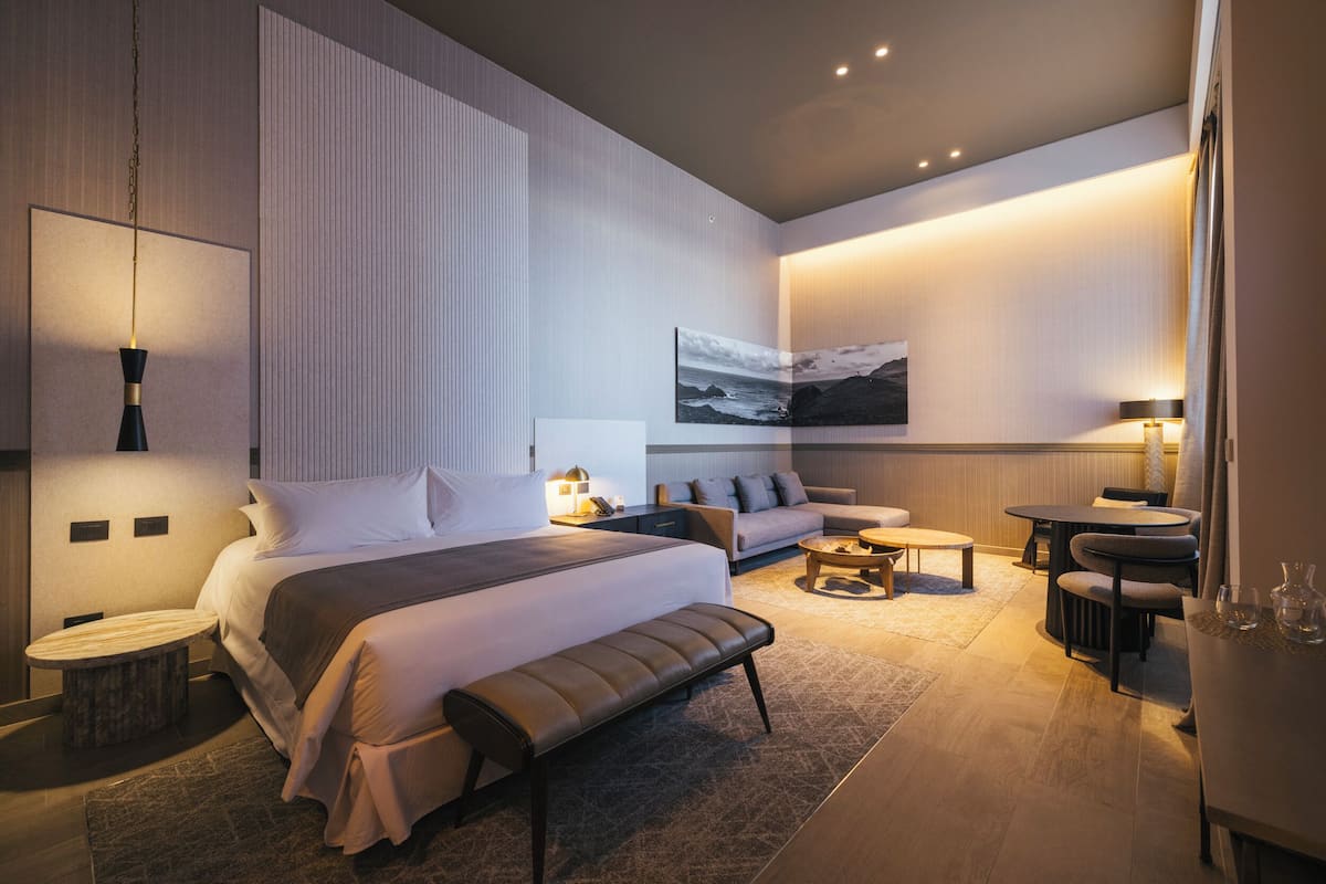 A modern hotel room with a large bed, a bench, a sofa, a round coffee table, and a dining area with chairs and a table.