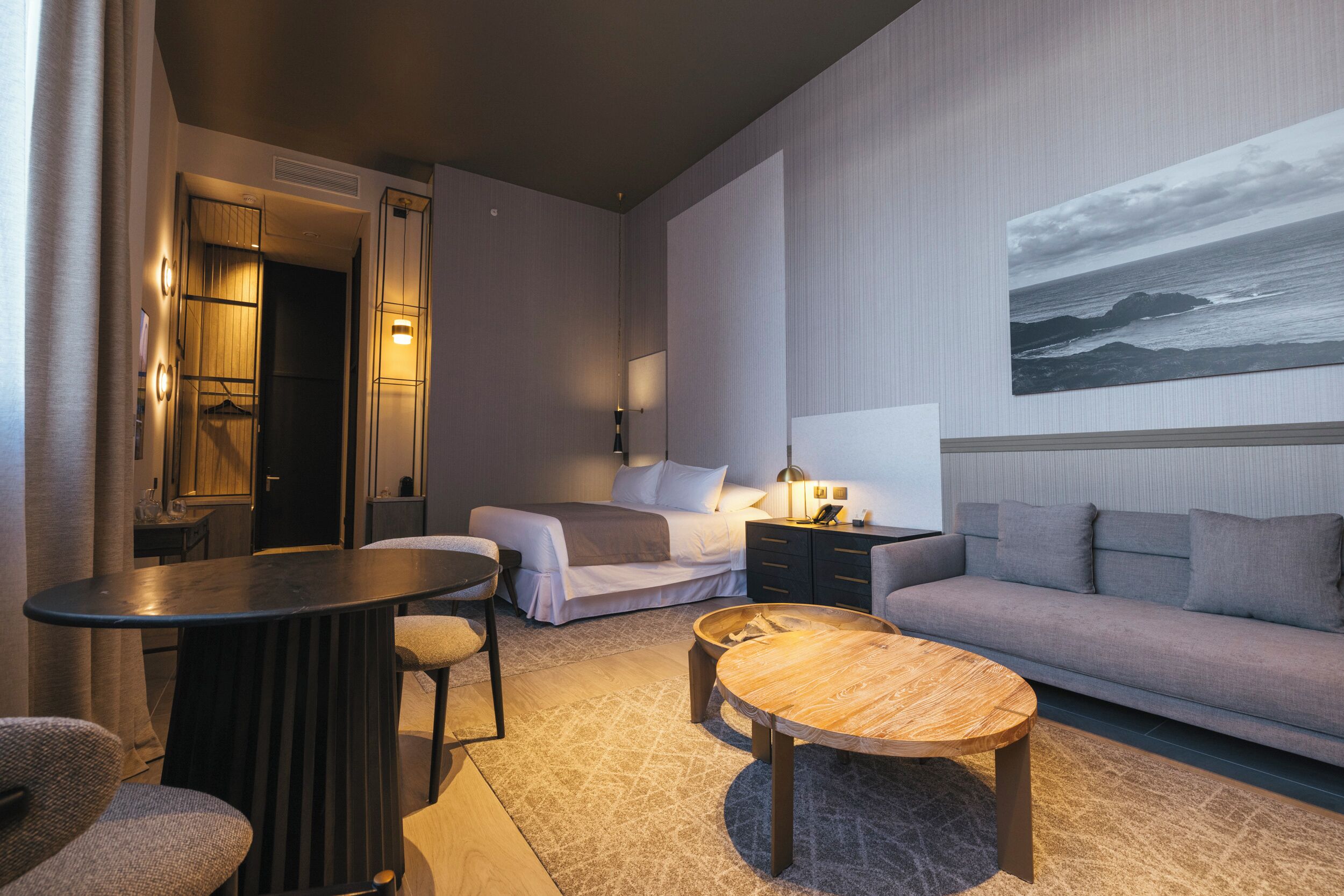 A modern hotel room with a bed, a sofa, a round wooden coffee table, a small round table, and a wall-mounted TV.