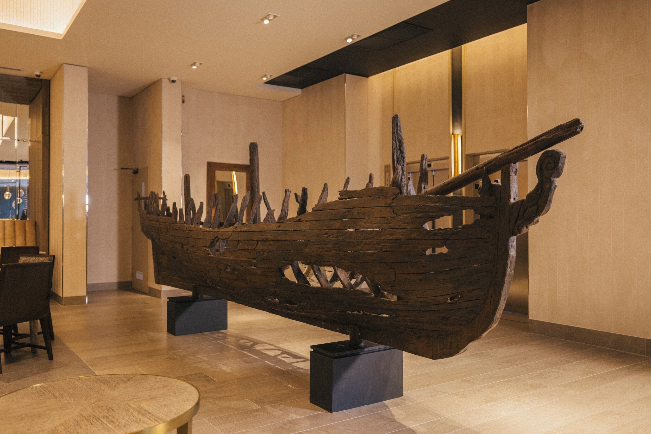 Une grande sculpture de bateau en bois est exposée à l’intérieur, dans un espace au décor moderne, avec des sols carrelés et un mobilier minimaliste.