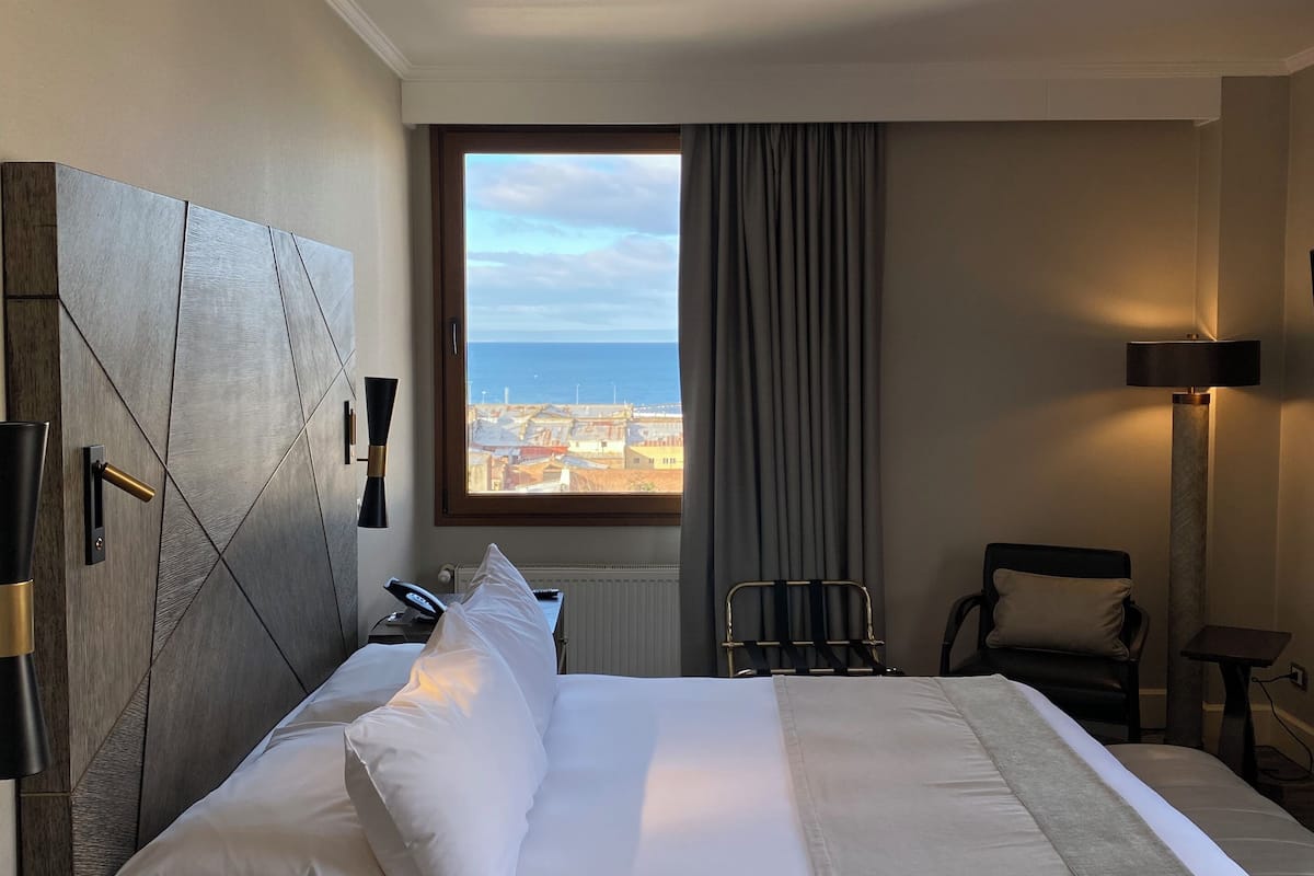 A hotel room with a bed, a view of the sea, a chair, a lamp, and a desk.