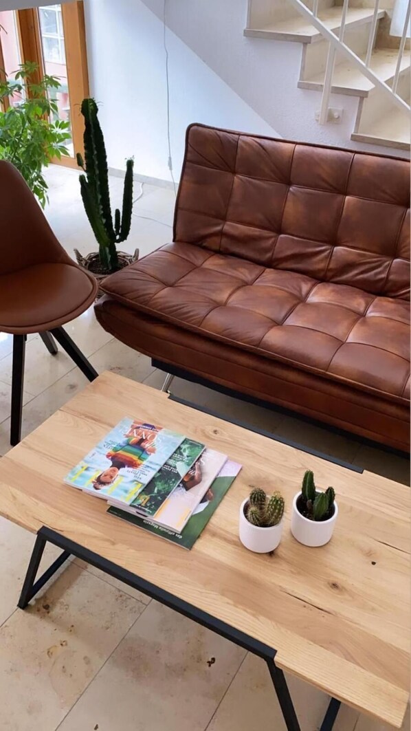 A brown leather sofa, a wooden coffee table with magazines and two potted cacti, and a chair in a room with a staircase in the background.
