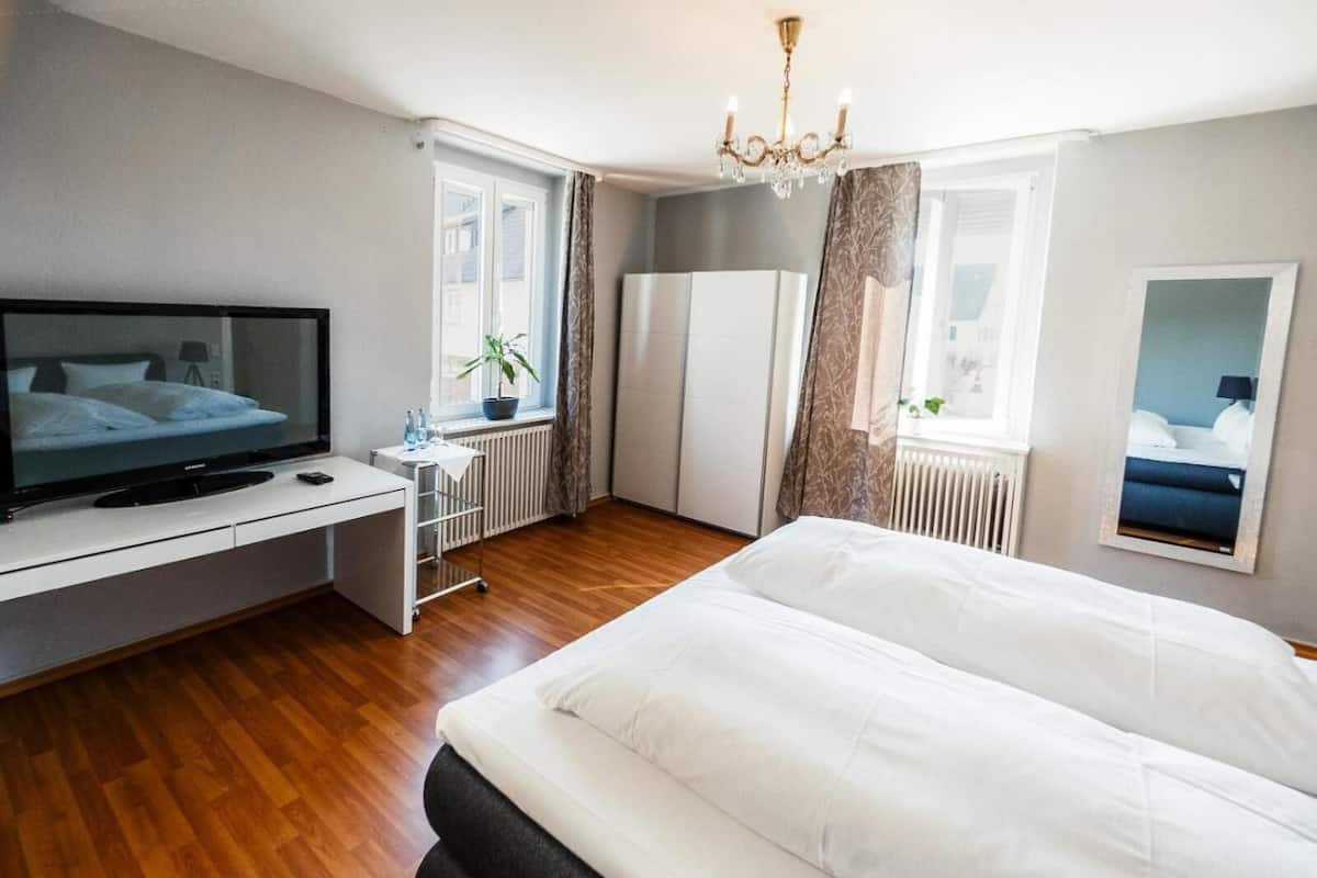 A bedroom with a bed, a TV, a desk, a radiator, and a window with curtains.