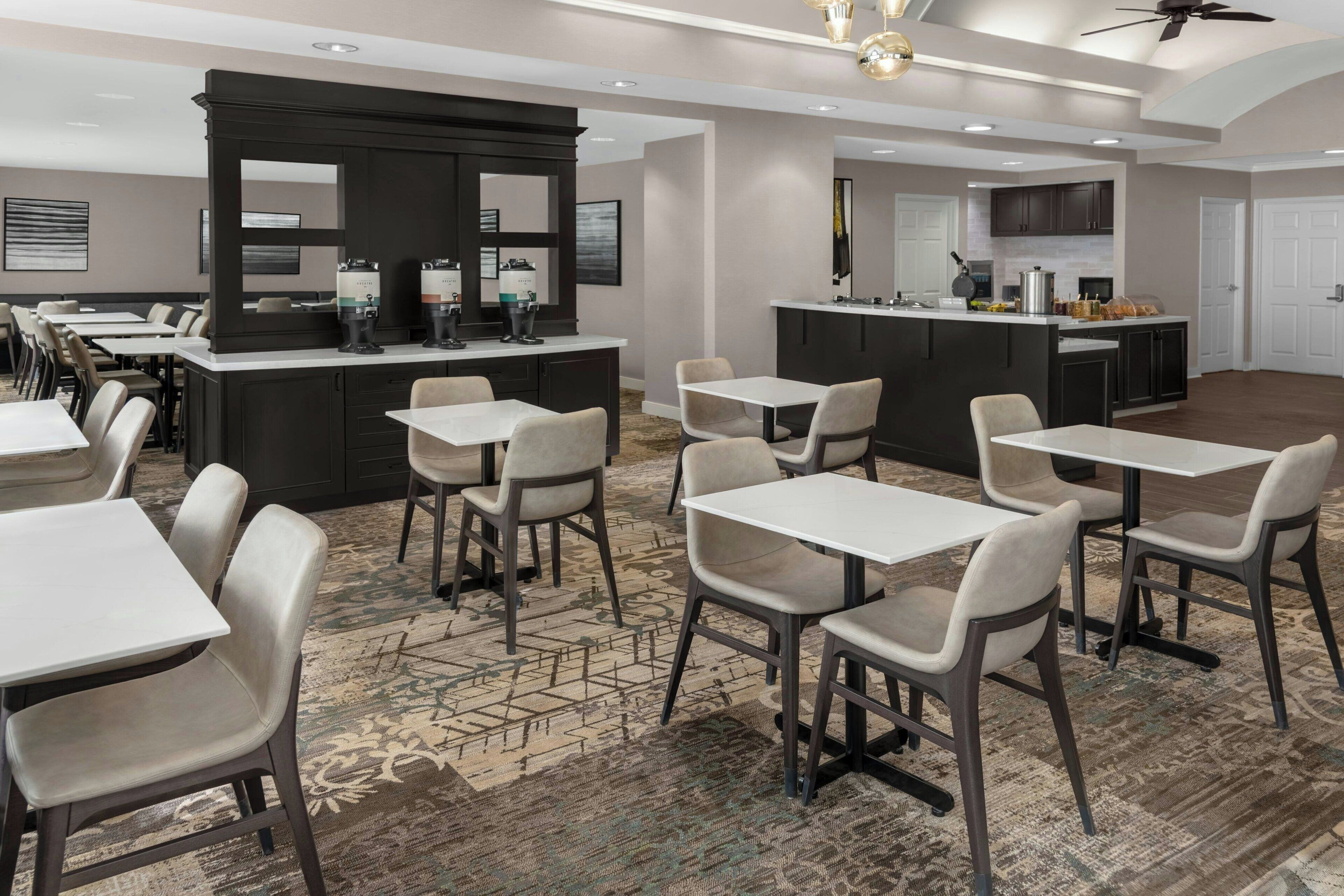 A modern hotel dining area with a bar, multiple tables and chairs, and a kitchen in the background.