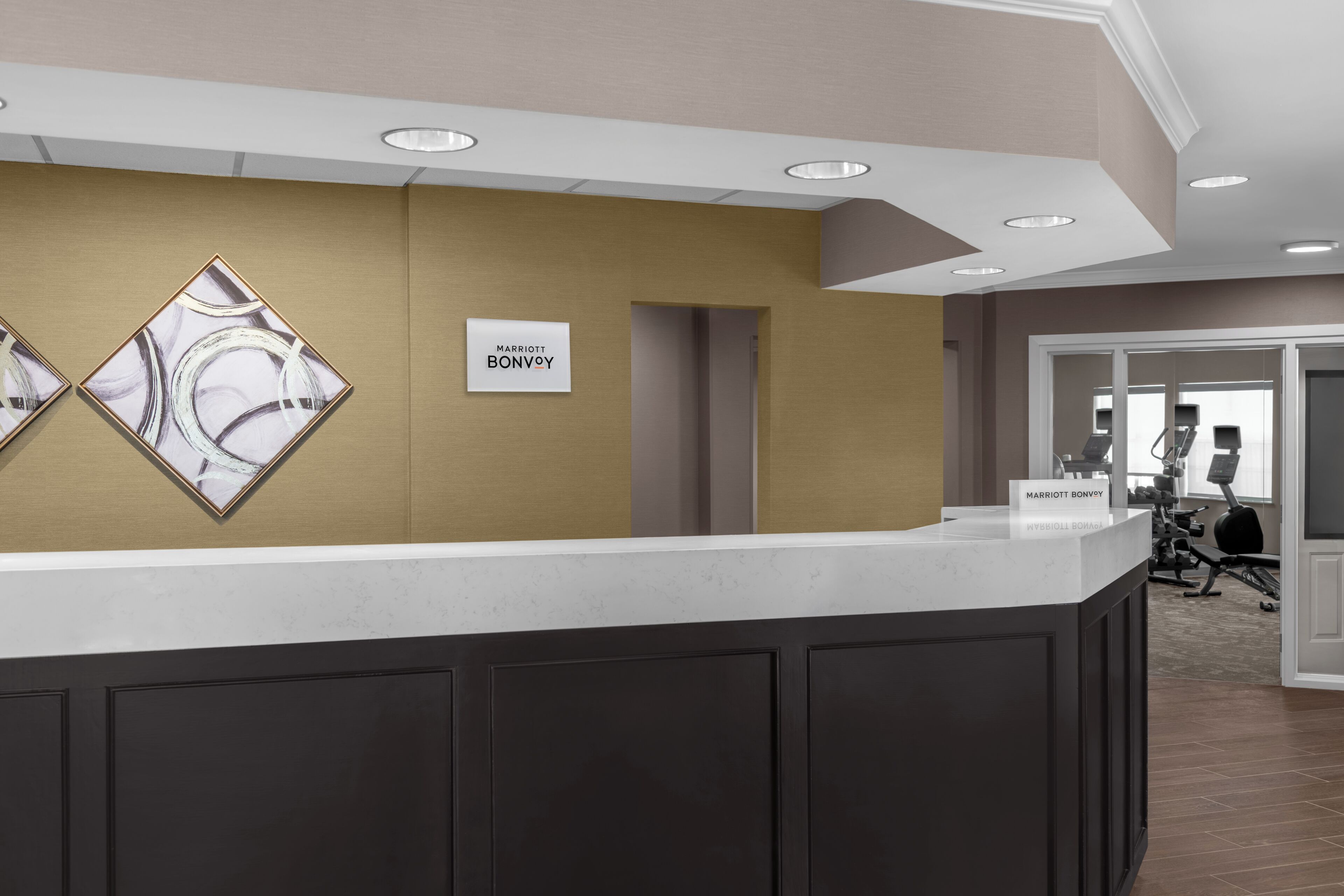 A hotel reception desk with a marble wall, a sign reading 'MARRIOTT BONVOY', and a glimpse of the gym area in the background.