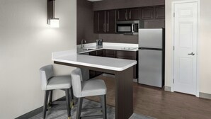 Une cuisine moderne avec des meubles de couleur sombre, un réfrigérateur en acier inoxydable, un four à micro-ondes et un plan de travail blanc. On y trouve deux tabourets de bar gris et un petit îlot central.