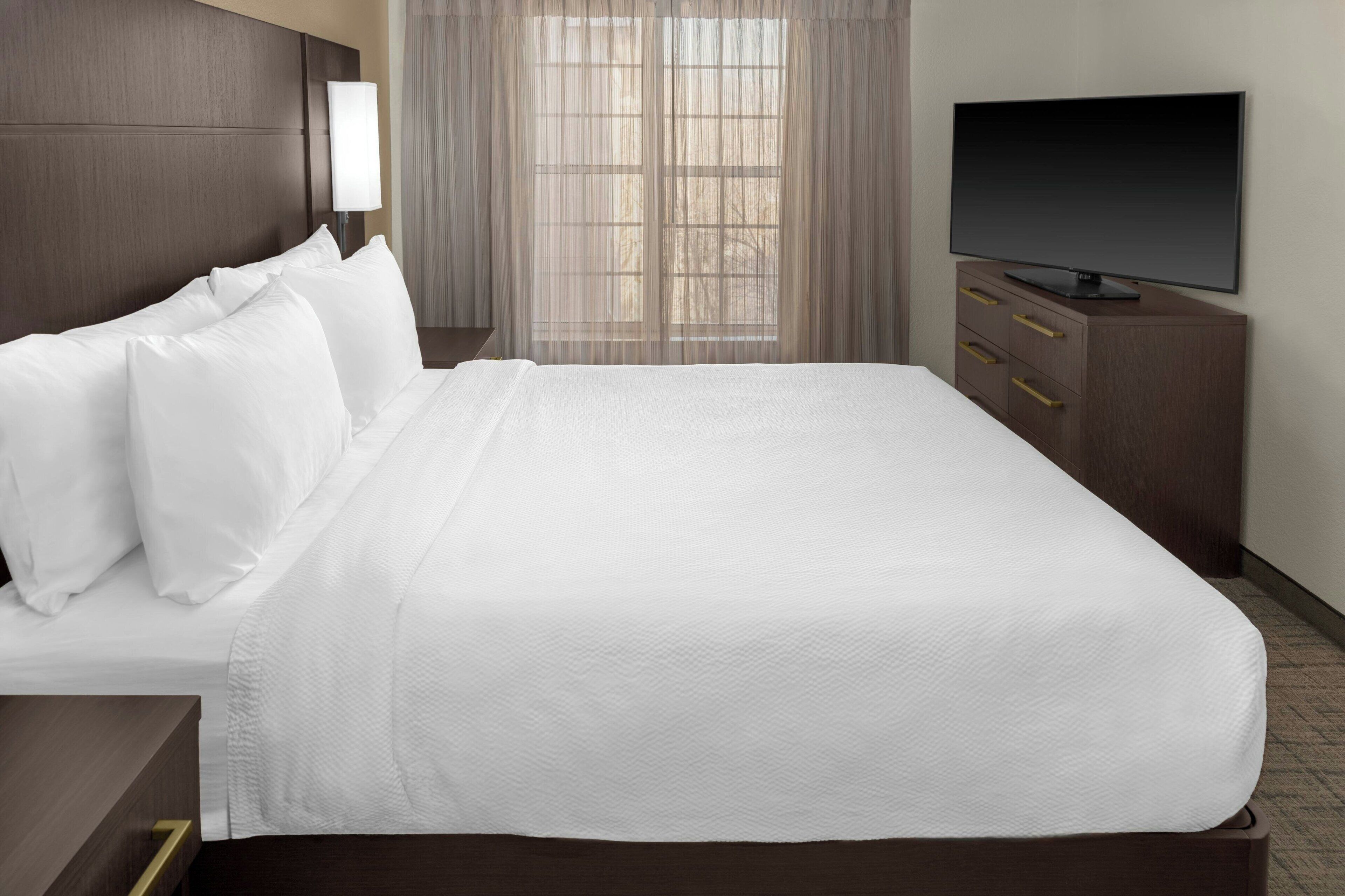 A neatly made bed with white linens, a television on a wooden cabinet, and a window with sheer curtains.