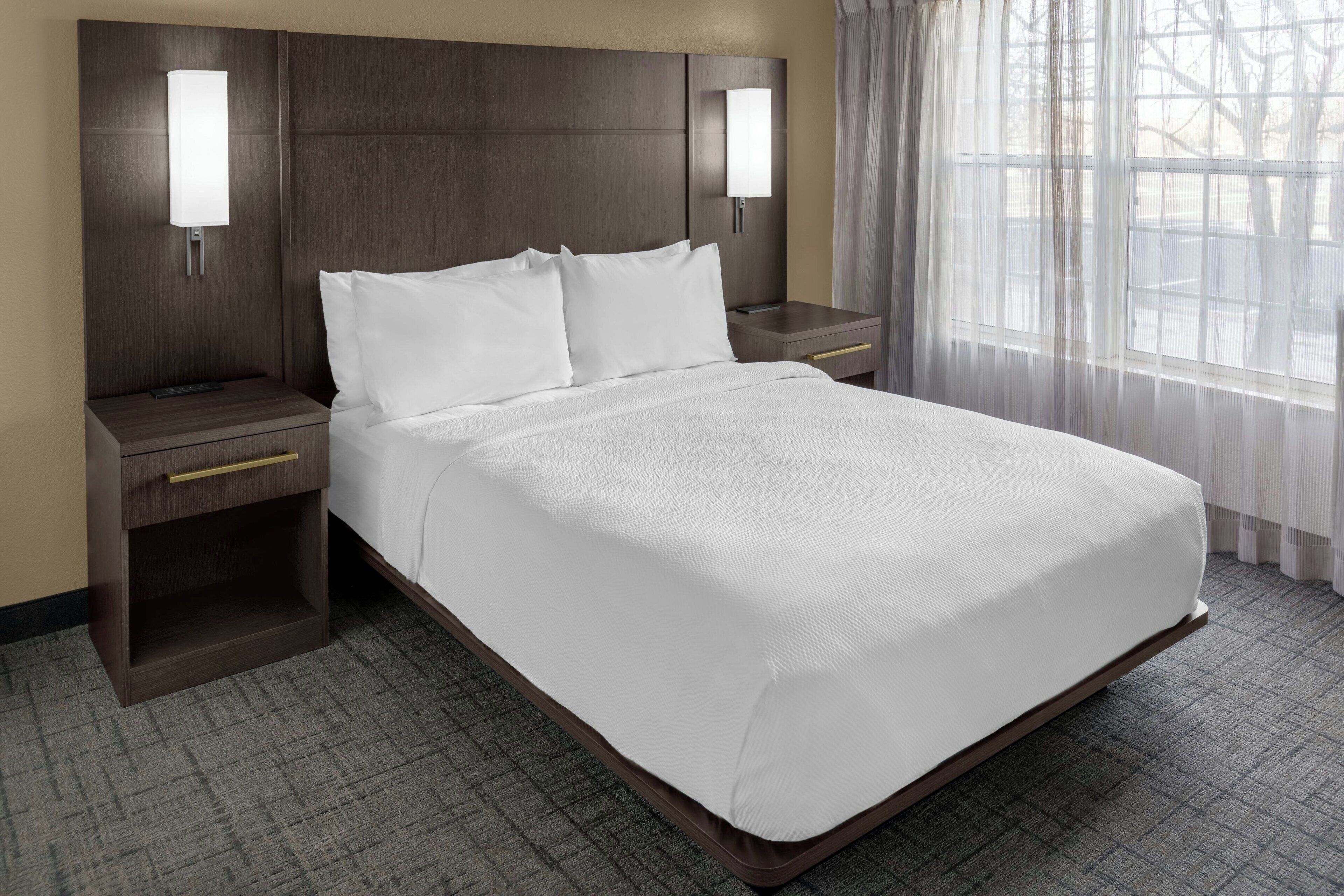 A hotel room with a large bed, two bedside tables, a nightstand, and a window with sheer curtains.