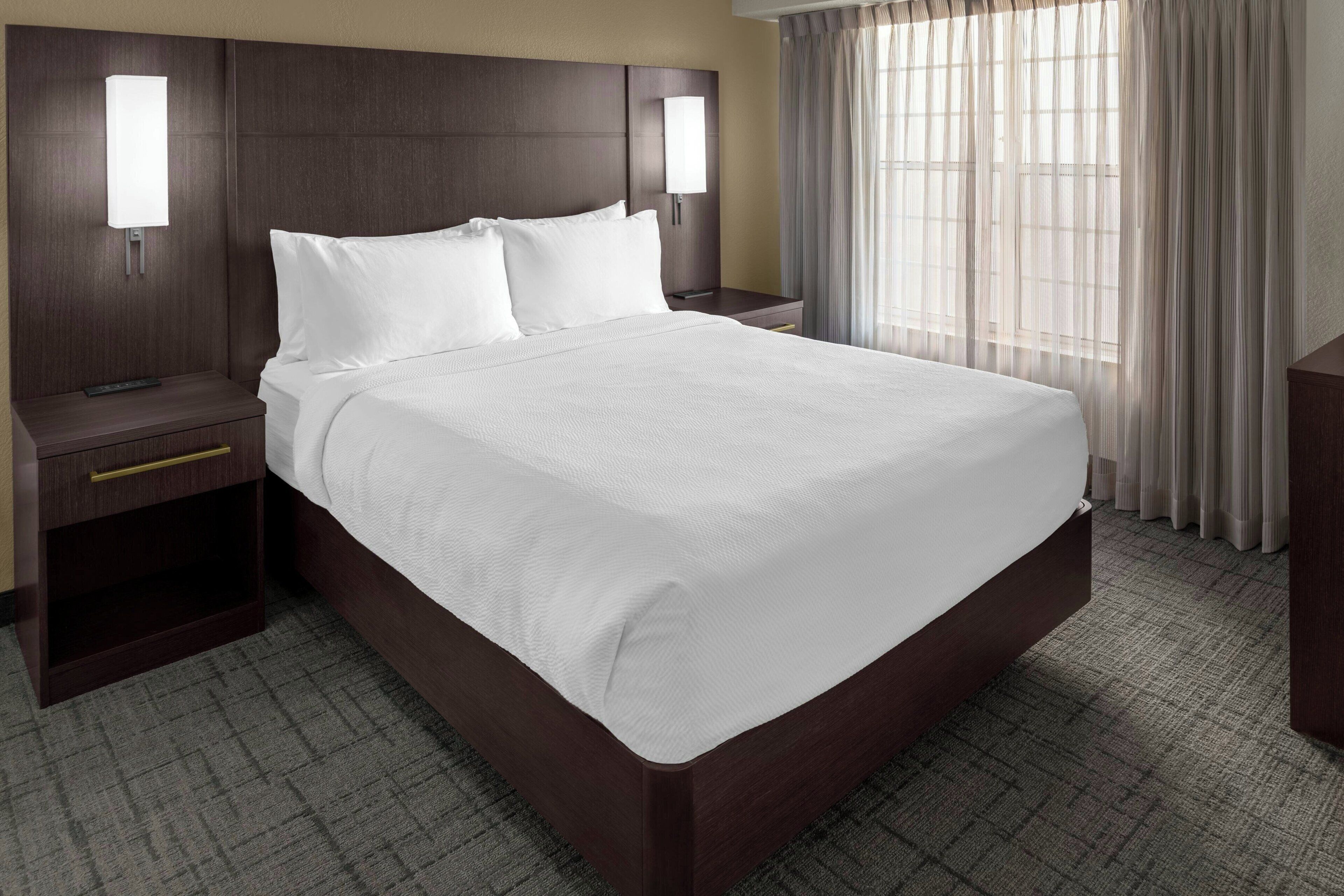 A hotel room with a large bed, two bedside tables, a nightstand, a lamp, and a window with curtains.