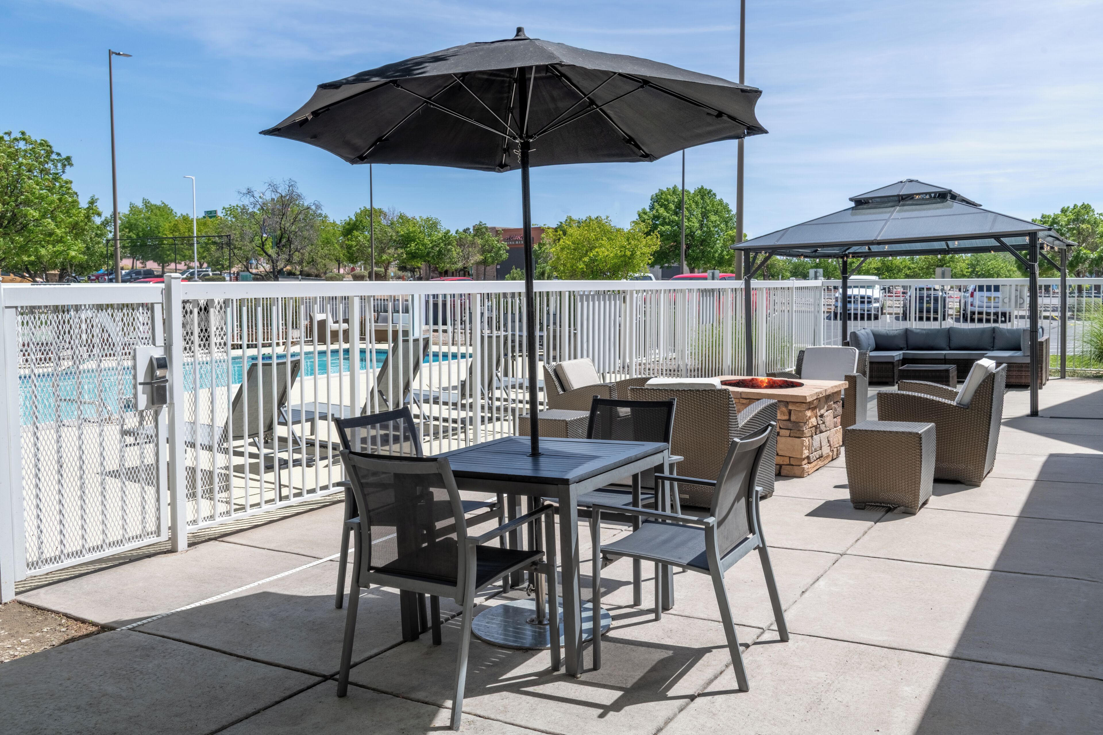 An outdoor patio with a pool, enclosed by a white fence, featuring a table and chairs, a grill, and a covered seating area.