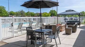 An outdoor patio with a pool, enclosed by a white fence, featuring a table and chairs, a grill, and a covered seating area.